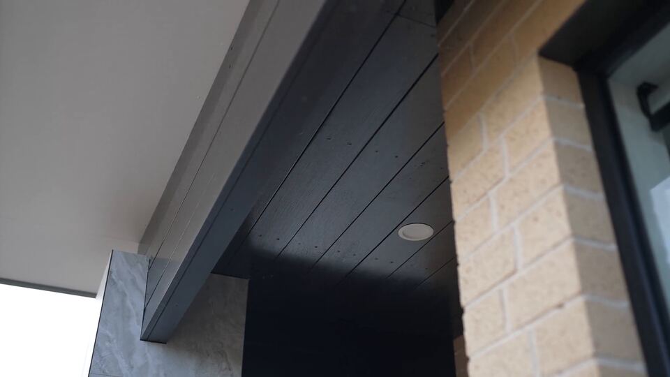 Porch detail where dark timber-look ceiling cladding meets cream brick and a black window frame